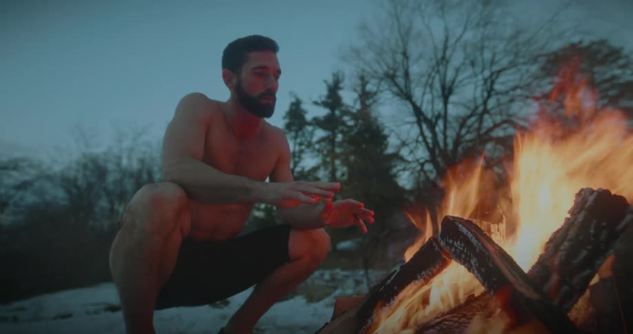 Man with no shirt and just shorts in a snowy forest on a foggy day warming himself in front of a campfire