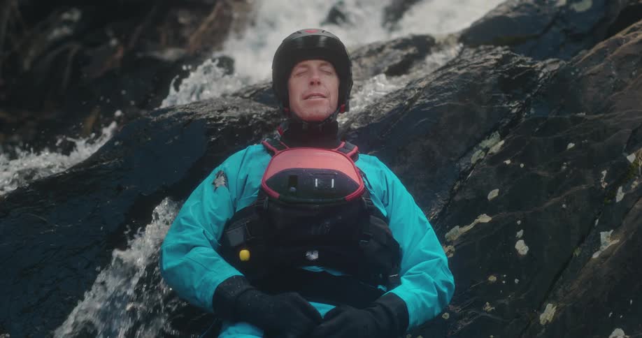 A man smiles slightly with his eyes closed as he sits in a river rapid