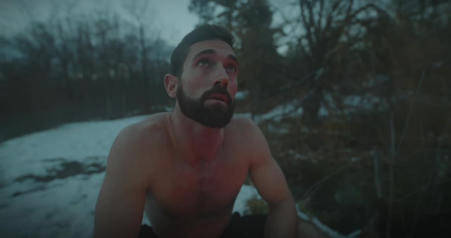 Portrait of a man crouching shirtless in a natural setting in the snowy forest warming himself with a campfire