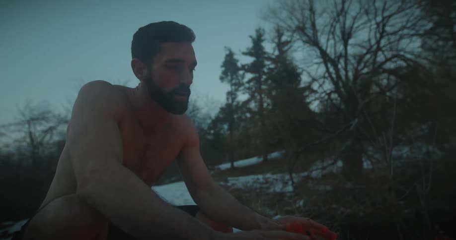 Man climbing out of a lake and warming his body in front of a bonfire in a snowy forest