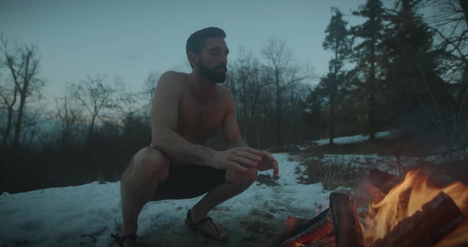 Shirtless man warms himself by a fire during winter