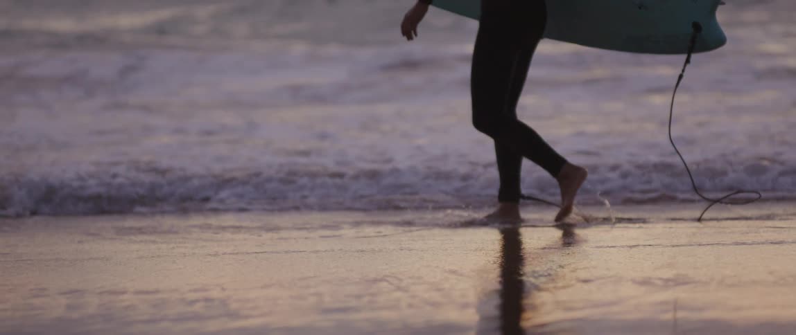 Surfer carrying surfboard attached to ankle with a leash walks through water on the beach