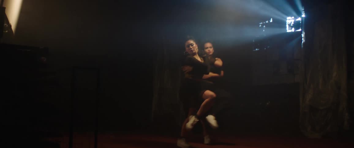 Two women are dancing choreographically in a dark room with a small source of natural light