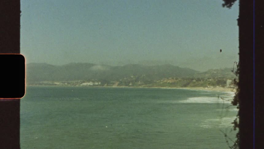 An old image of a sea with clear water next to a beach with lots of sand and in the background there are mountains