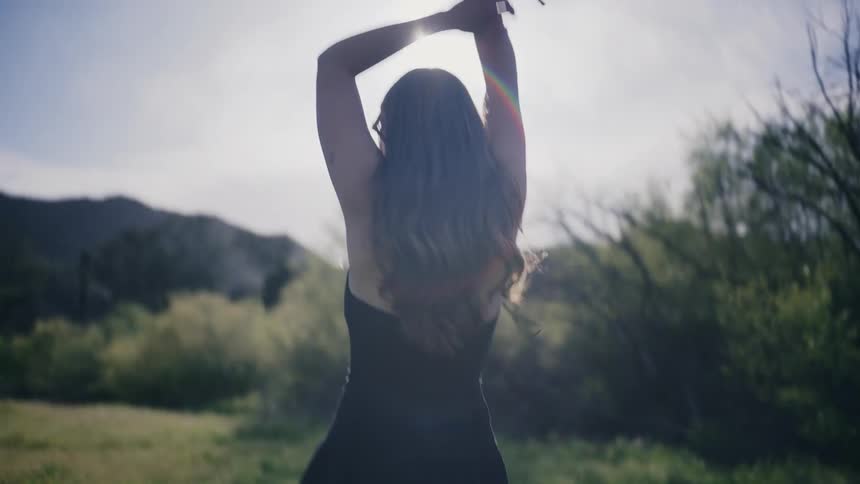 A young woman in a black dress holding flowers walks through a field lifting her arms up