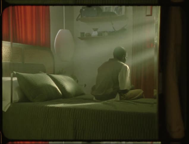 A man sits up on a bed bathed in flickering sunlight