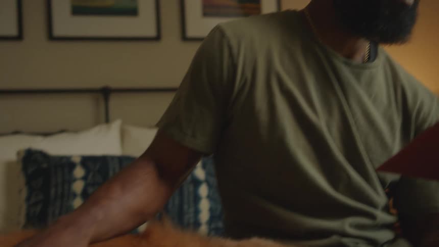 Man sitting on the bed reading a book while massaging his dog who is lying next to him