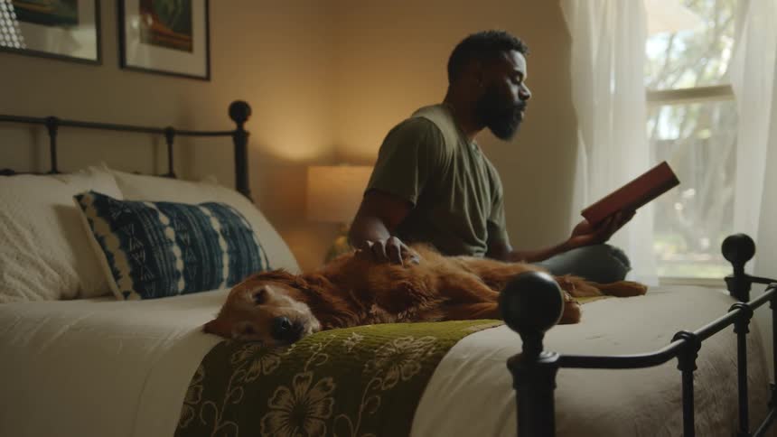 A black man sitting on a bed reads a book while petting his dog