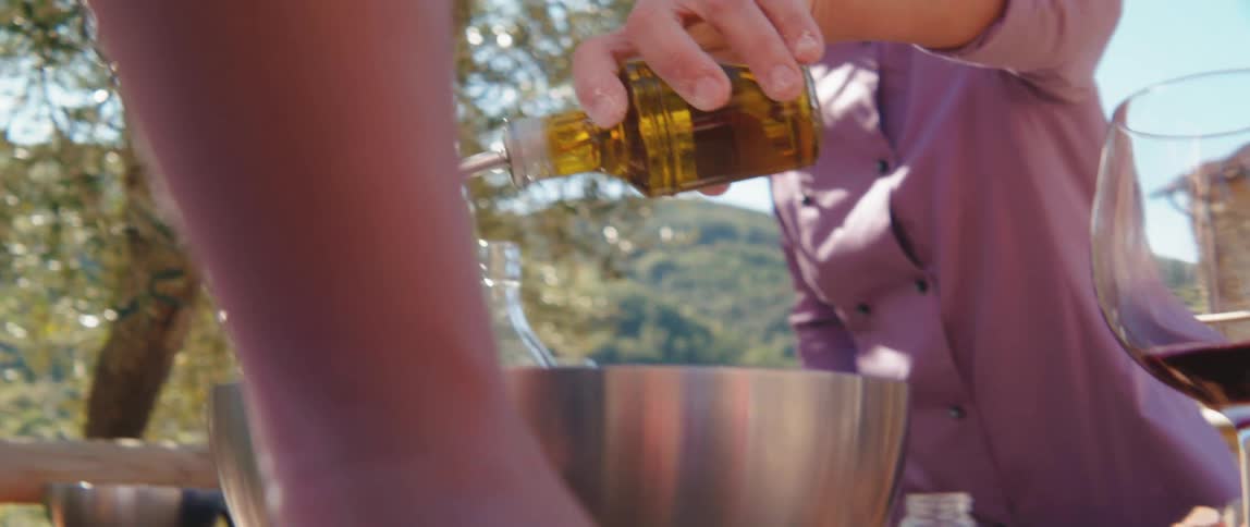 Man drizzles oil into metal bowl sitting on picnic table in yard of house near a mountain