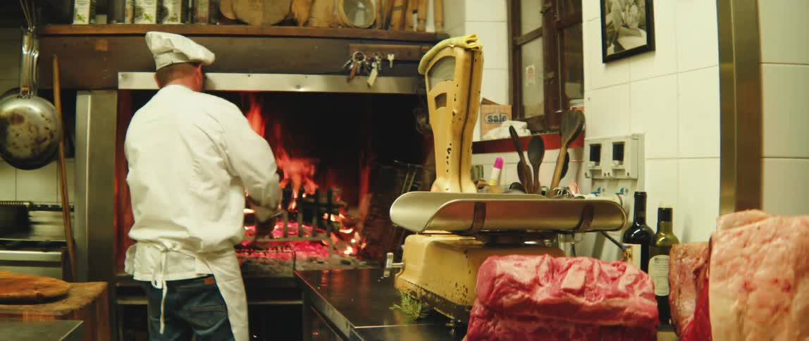 Man in an apron placing coals on a grill in a restaurant kitchen with raw meats on the counters around