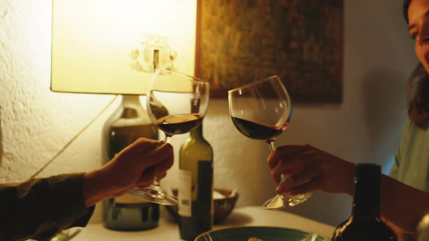 Hands of a couple sitting at a table with plates of pasta and clinking wine glasses