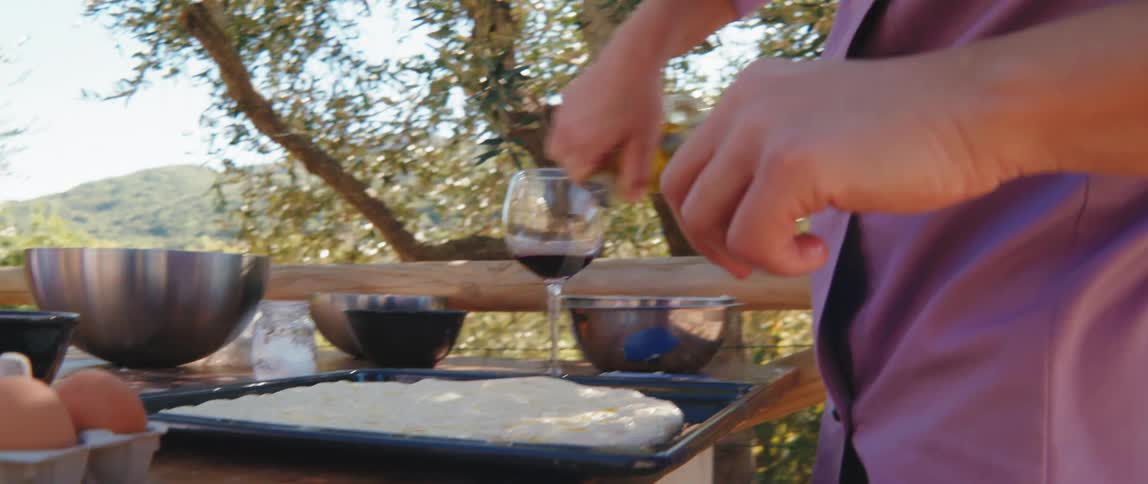 A womans hands pouring olive oil into a dough that is on a wooden table outdoors
