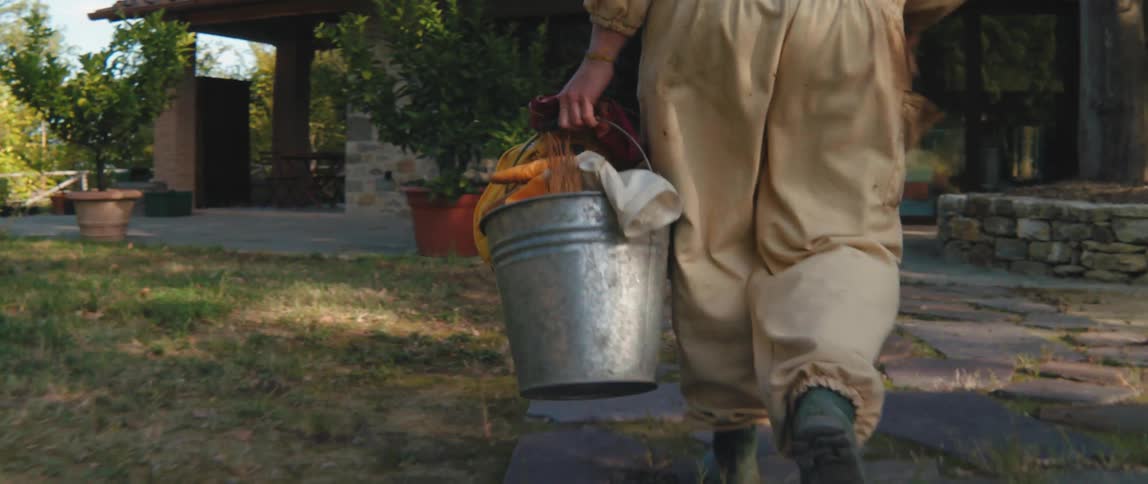 A person in beige clothing carrying a metal bucket walking on stone slabs