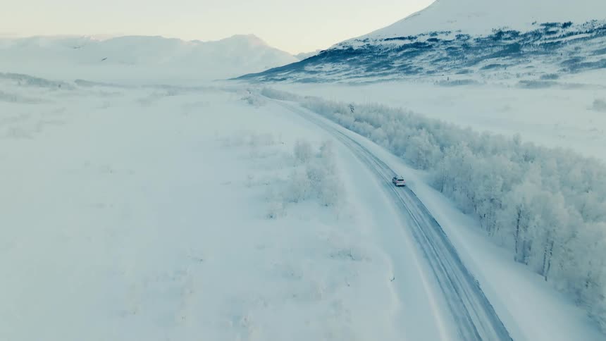 Aerial of a car driving on a snowy road near the snowcovered trees and mountains