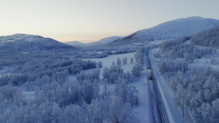 Aerial view of cars and trucks driving on road in the snowy field near the forest at sunset