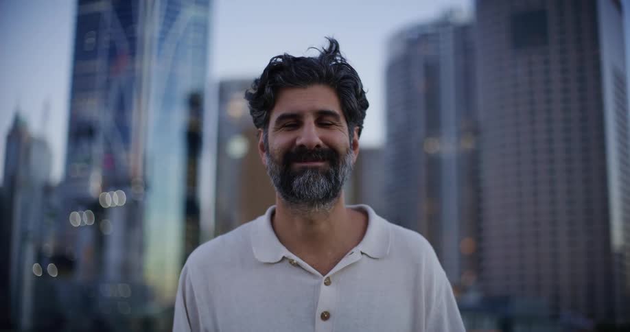 The face of a man who is happy and he has a beard on his face and is wearing a white shirt and behind him there are several buildings in blur