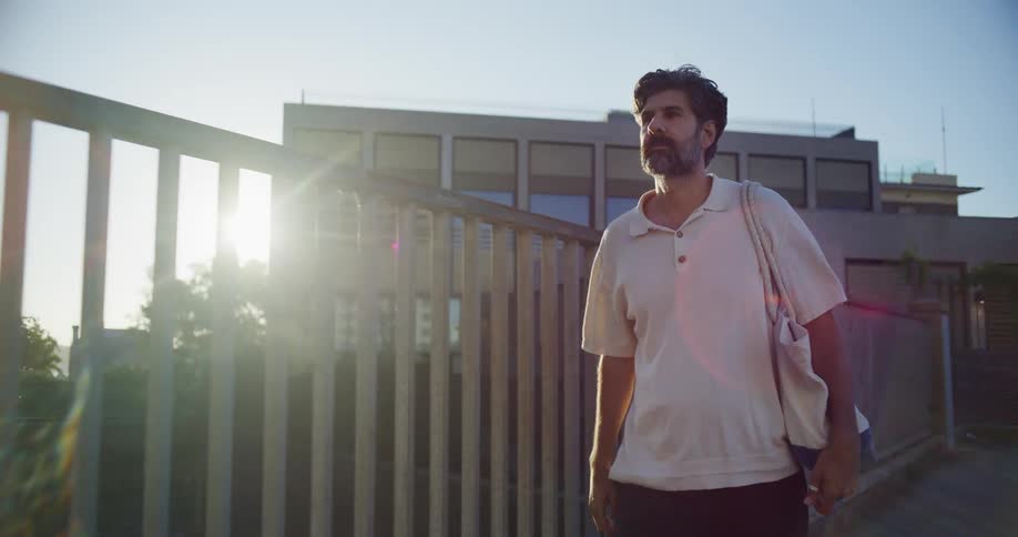 Man carrying a bag walks along urban path as sun shines past a building in the background