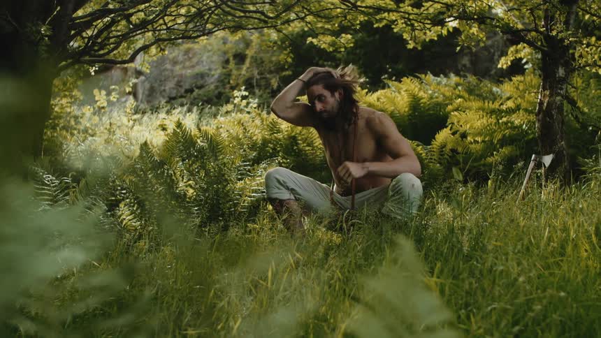A shirtless individual seated on the ground among tall grass