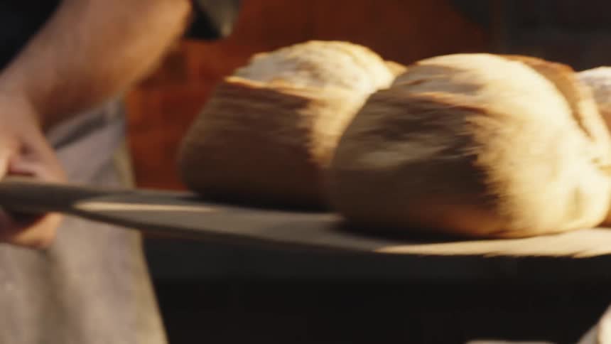 Hands removing fresh bread from a woodfired oven with a wooden paddle