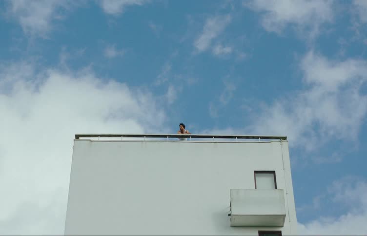 A person is on the terrace of a white building observing the sides and the sky is blue with some white clouds passing quickly