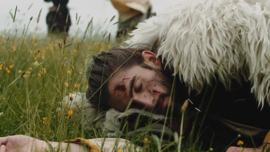 Portrait of a viking man falling to the grass with a bloody face and other warriors fighting around him