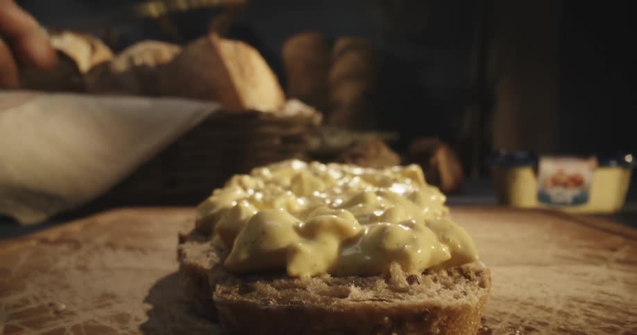 A persons hand with a knife applying sauce to a slice of bread on a wooden table
