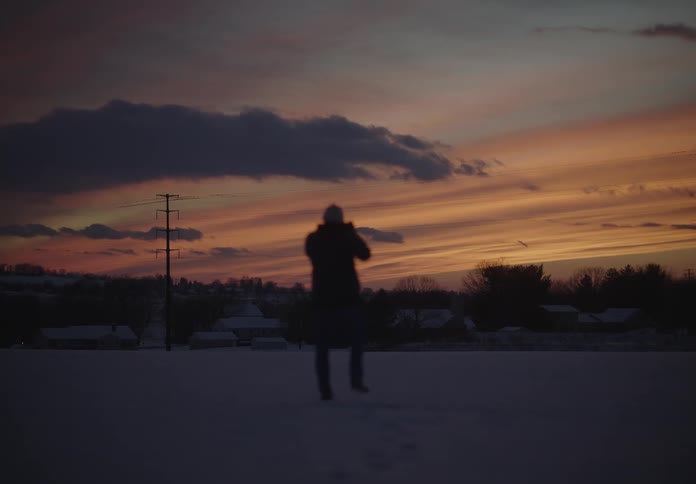 A person has his back turned and is walking on snow with the orange sky in the morning