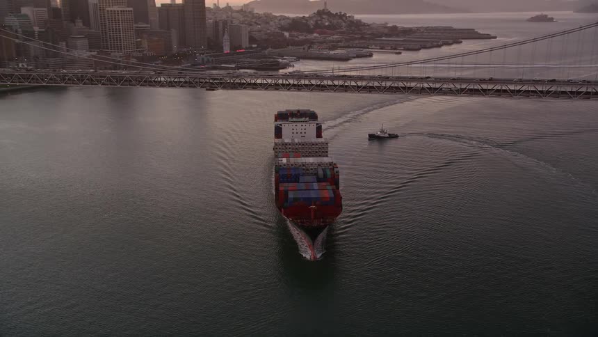 Aerial panorama of a cargo ship with containers passing under a bridge and in the background a metropolis with a skyscraper in the morning