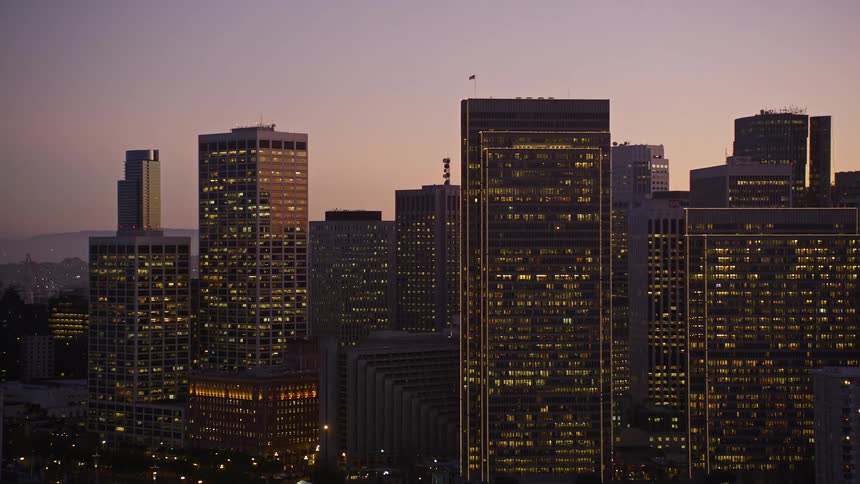 Interior lights shine from the windows of skyscrapers in a city stretching between the water and the mountains