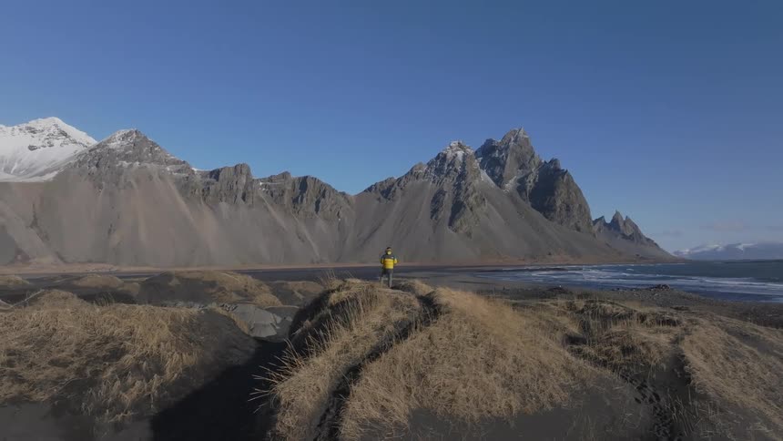 A person is in a yellow jacket standing on a hill looking at a sea coast with several mountains on the side