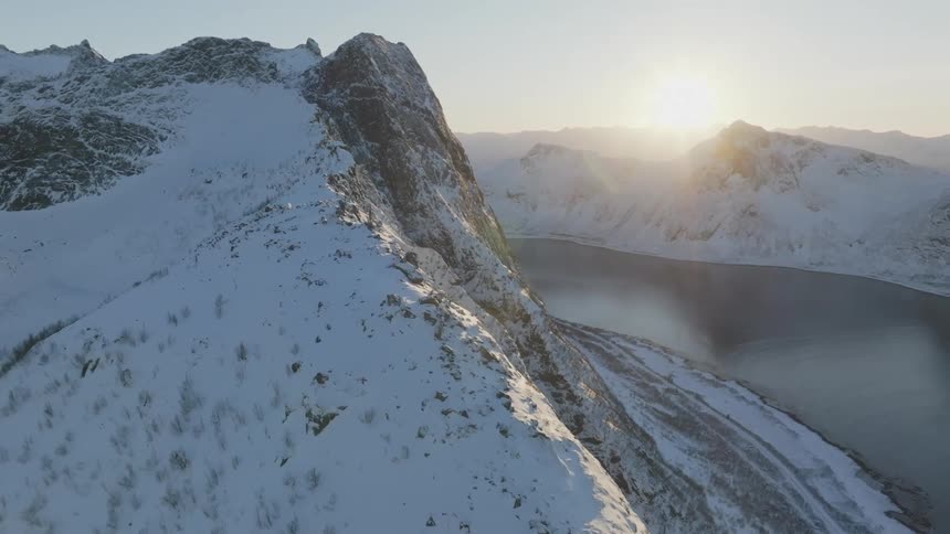 Aerial panorama of a mountain with a snowcovered cliff and a river lake around it with the sunrise on the horizon