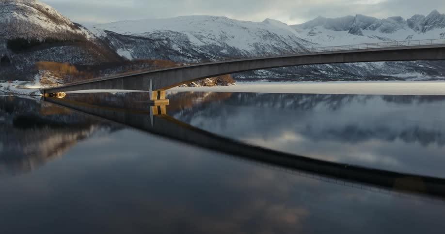 A suspension bridge on a still lake that is reflecting the image of the bridge in a region with many snowcovered mountains