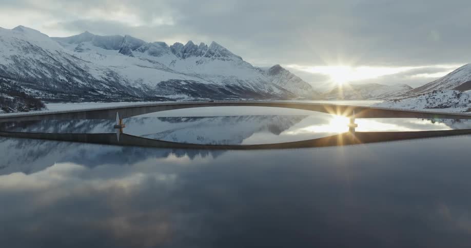 A long bridge extends over a still body of water reflecting the snow covered mountains at morning
