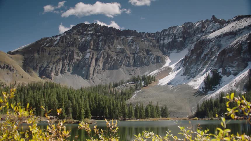 Deciduous trees line the shore across the lake fromthe forest and mountains on a cloudy day
