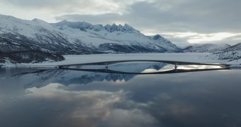 A suspension bridge over a still lake that is reflecting the image of the bridge in an environment with snowcovered mountains