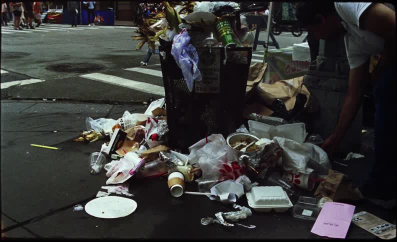 Person puts empty food containers on the ground close to overflowing trashcan near city street