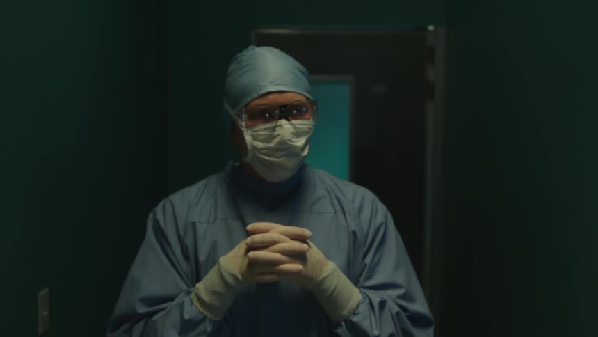 A surgeon walks through a doorway in a hospital and unclasps his hands