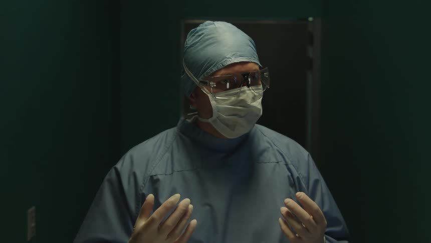 A doctor wearing surgical attire enters a dimly lit hospital room