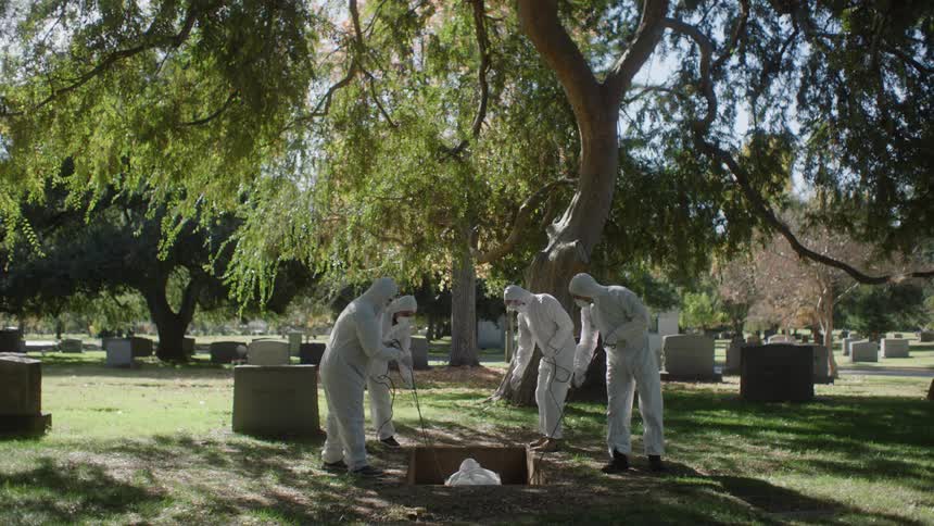 A group of four people in white anticontamination suits are burying a body in a hole in a cemetery
