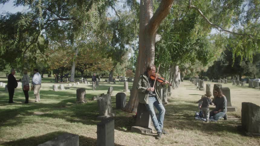 A man plays a fiddle in a cemetery as a funeral comes to a close