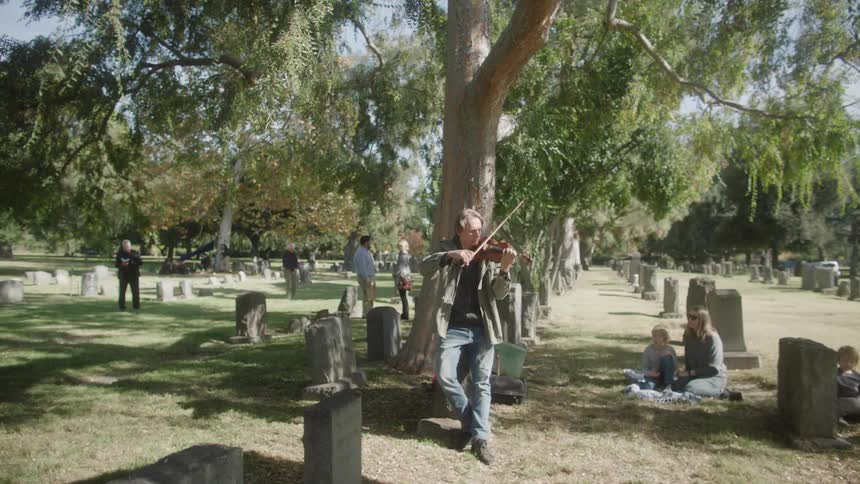 Musician leaning against a tree playing a violin in the middle of a cemetery with people around