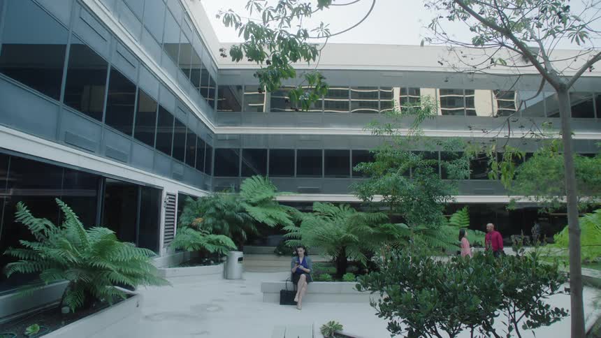 A woman is sitting outside a building with lots of vegetation and she is using a cell phone