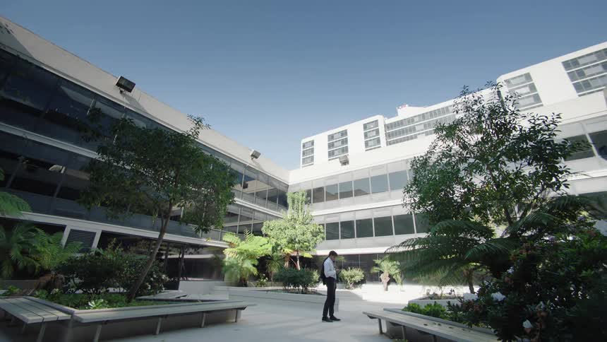 A man is standing outside a white building and there are several trees and vegetation