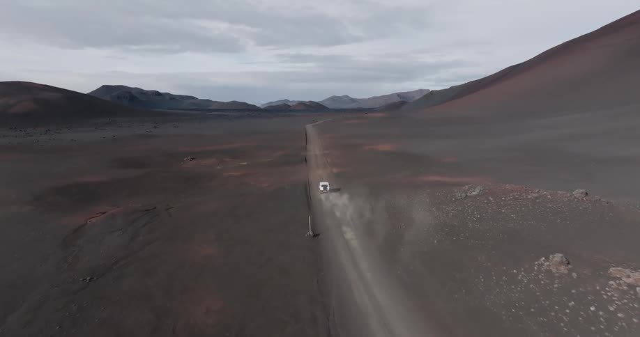 Aerial panorama of a white vehicle traveling on a dirt road in a desert environment with mountains around it on a cloudy day