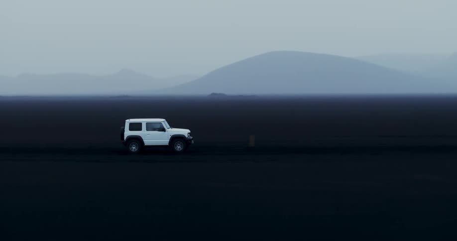 White jeep car traveling on black ground with silhouettes of mountains on the horizon on a foggy day