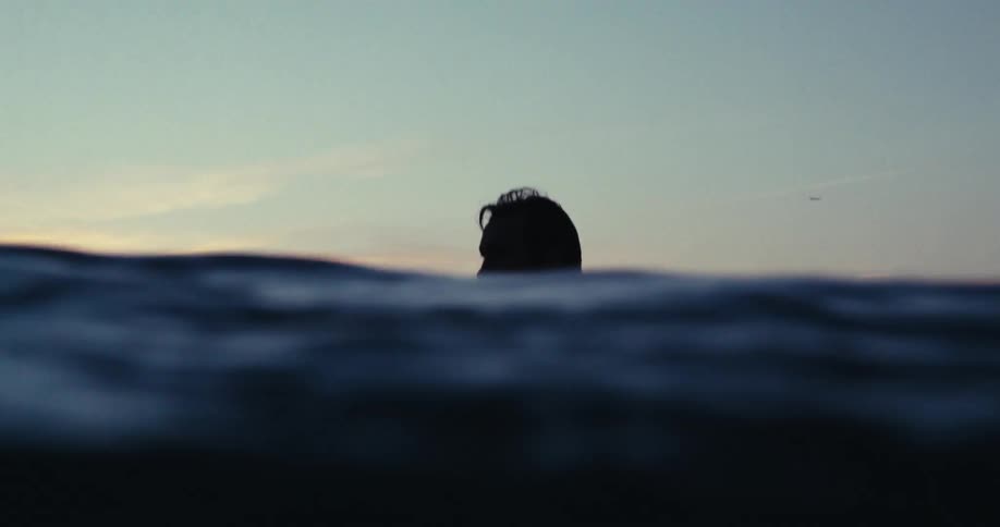 Man partially silhouetted against a red tinted horizon bobs in the ocean
