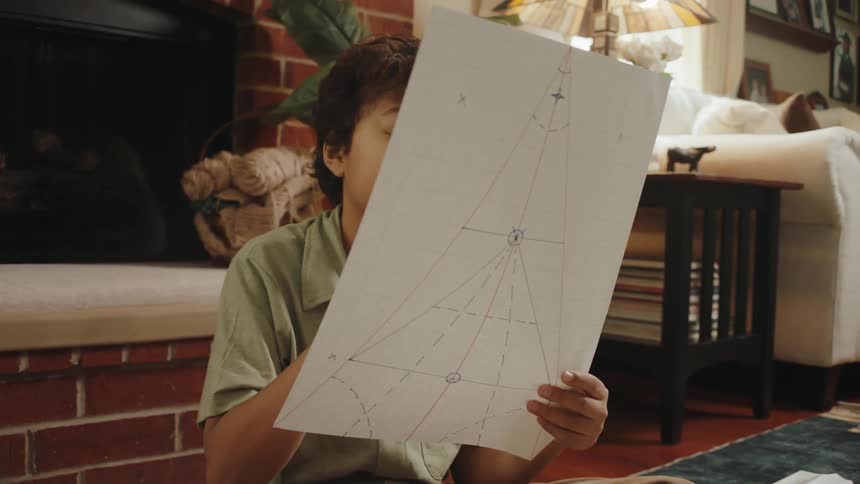 A boy sitting crosslegged on the floor going through large sheets of paper spread around him