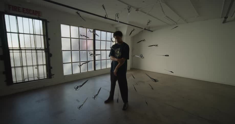 A young man surrounded by floating scissors on a room next to large windows
