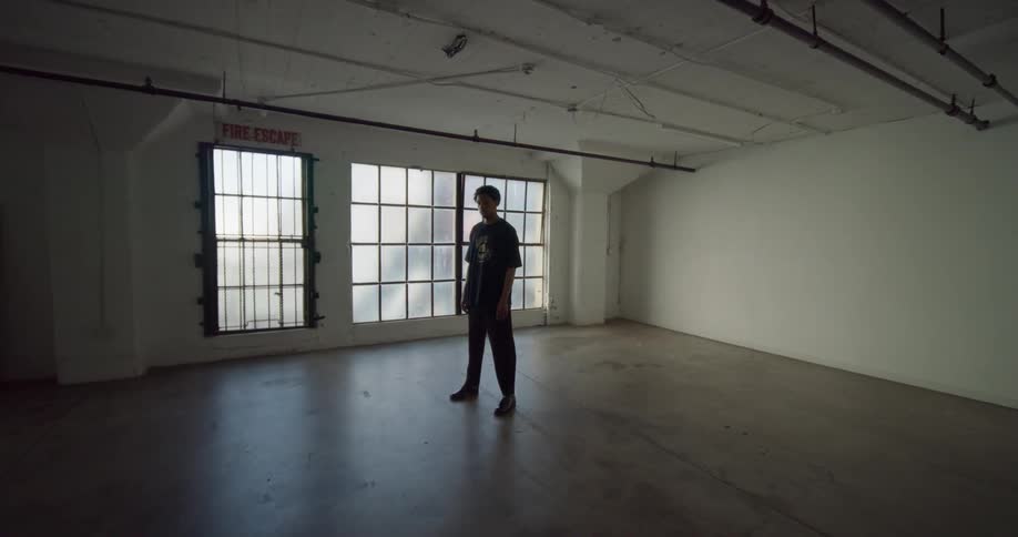 A young boy is standing in the middle of an empty room with large windows and is staring straight ahead