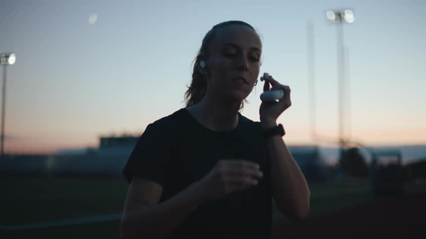A woman standing on a running field and she is putting headphones in her ears